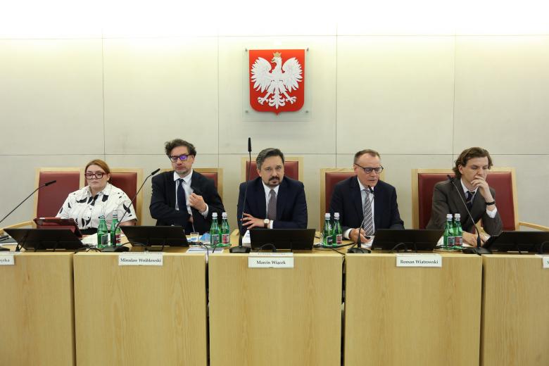 Pięć osób siedzi za stołem prezydialnym, na ścianie za nimi godło Polski - biały orzeł w koronie na czerwonym tle