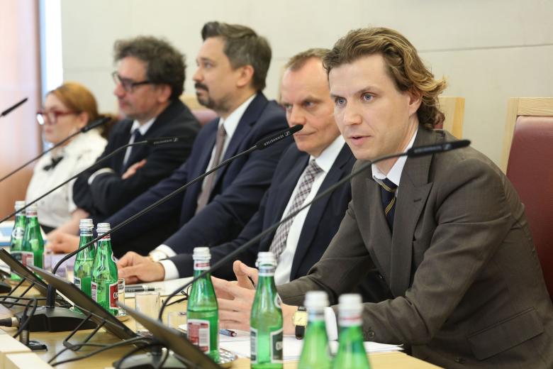 Pięć osób siedzi za stołem prezydialnym, osoba na pierwszym planie mówi do mikrofonu