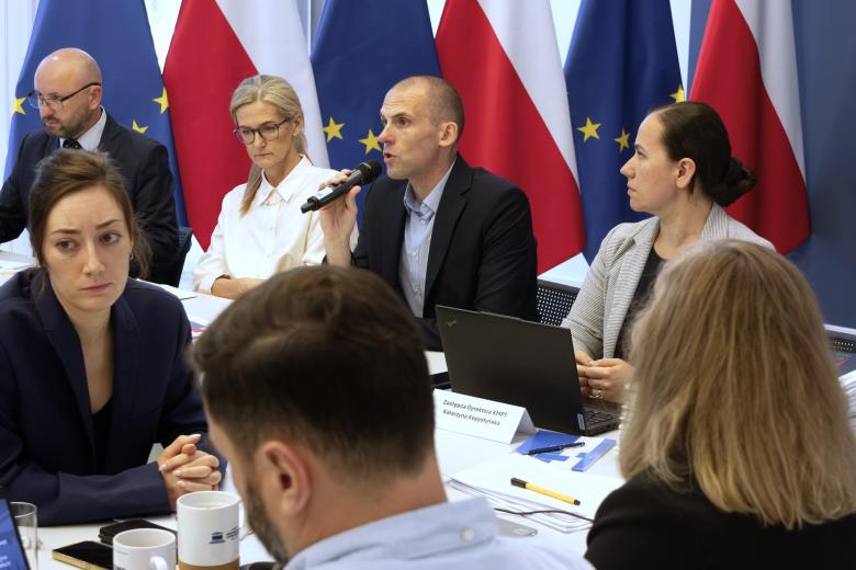 Osoby siedzą za stołem na tle flag Polski i UE, jedna z nich mówi do trzymanego w dłoni mikrofonu