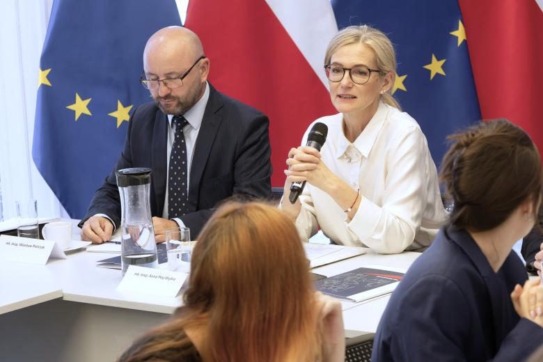 Osoba siedząca za stołem na tle flag Polski i UE mówi do trzymanego w dłoni mikrofonu
