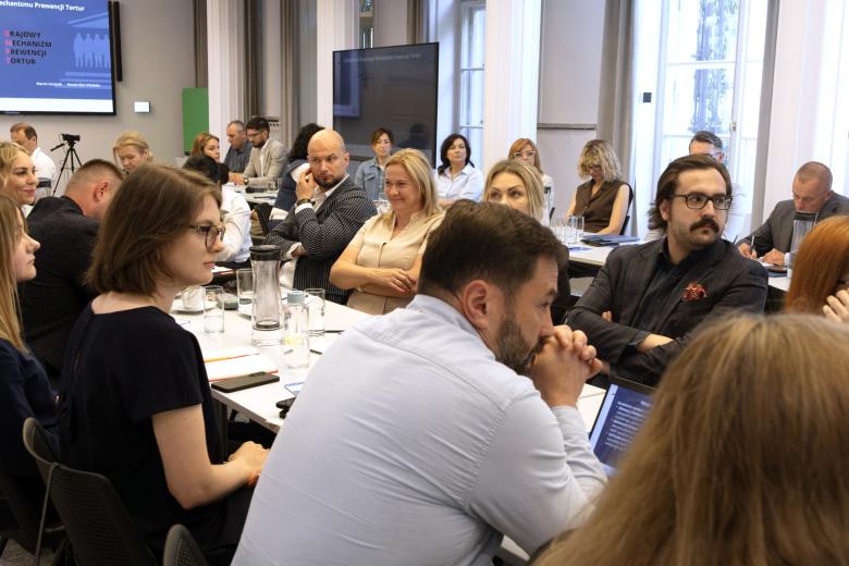Kilkadziesiąt osób siedzi przy stołach ustawionych w podkowę na dużej sali konferencyjnej