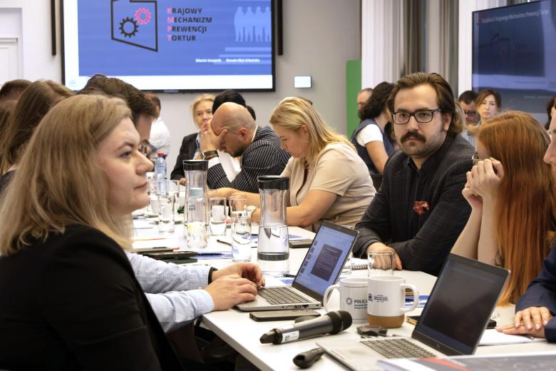 Kilkanaście osób siedzi przy stołach ustawionych w podkowę na sali konferencyjnej