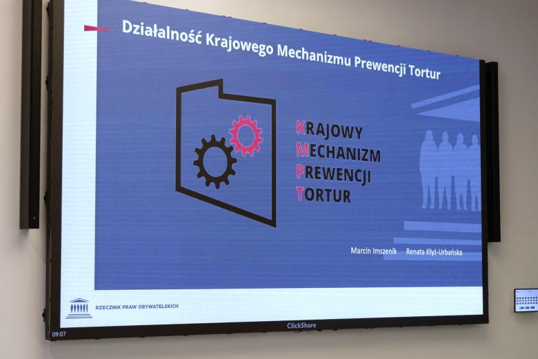 Duży ekran z wyświetlonym slajdem tytułowym prezentacji z napisem "Działalność Krajowego Mechanizmu Prewencji Tortur"