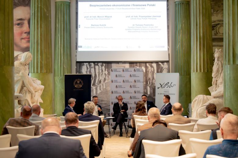 Pięć osób siedzi przy stolikach na sali konferencyjnej, przed nimi na widowni kilkadziesiąt osób siedzi i przysłuchuje się dyskusji