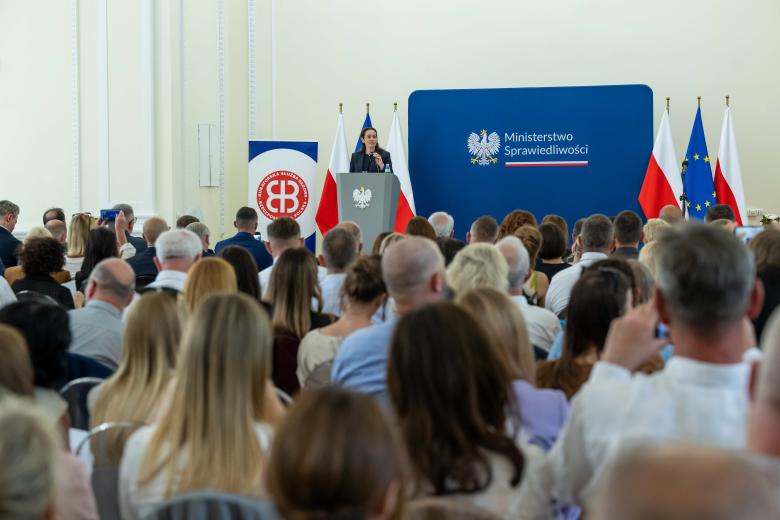 Kilkadziesiąt osób na dużej sali słucha wystąpienia osoby mówiącej za mównicą na tle planszy z napisem "Ministerstwo Sprawiedliwości"