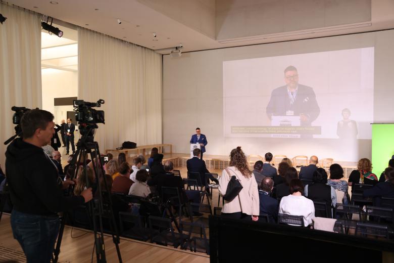 Widok sali konferencyjnej, w której siedzi kilkadziesiąt osób. Przed nimi przy mównicy stoi mężczyzna i wypowiada się.