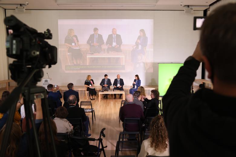 Cztery osoby siedzą przy stolikach naprzeciwko publiczności w sali konferencyjnej