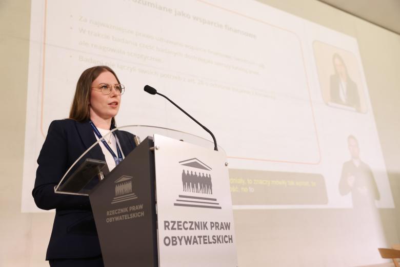 Angelika Greniuk wypowiada się stojąc przy mównicy. Za nią widoczna jest prezentacja
