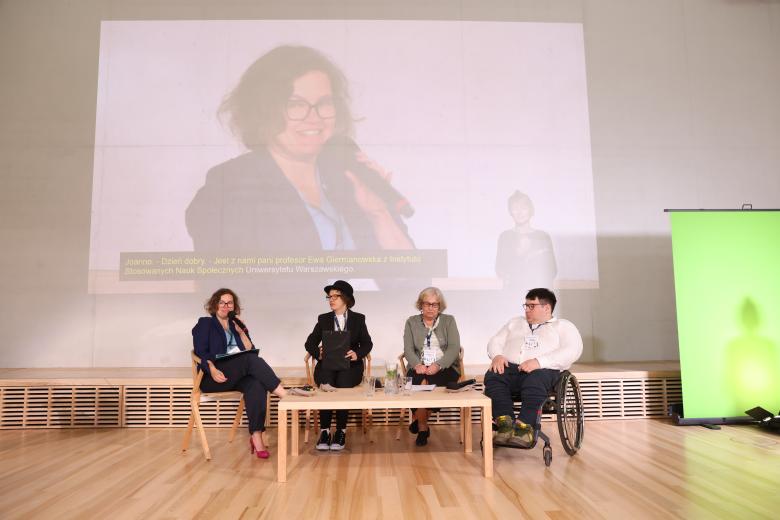 Cztery osoby rozmawiają siedząc przy panelu w sali konferencyjnej. Jedna z nich siedzi na wózku.