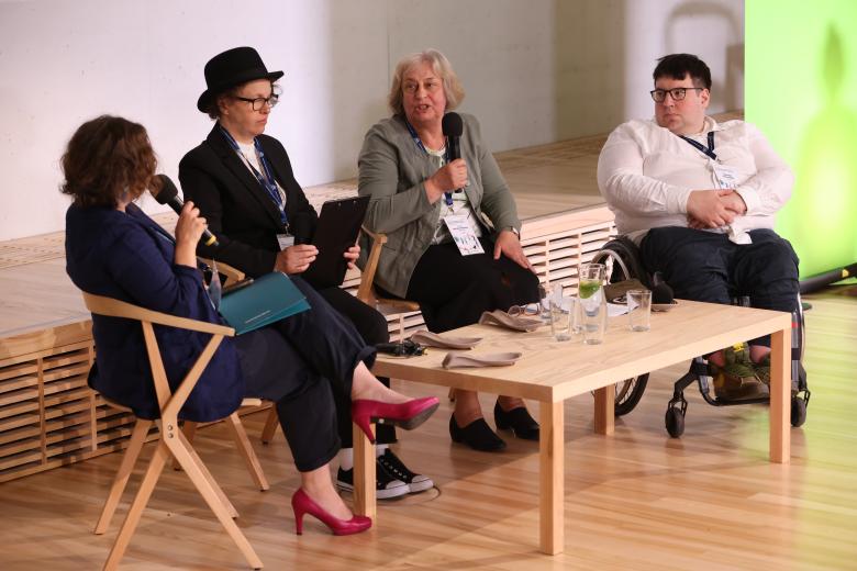 Cztery osoby rozmawiają siedząc przy panelu w sali konferencyjnej. Jedna z nich siedzi na wózku.