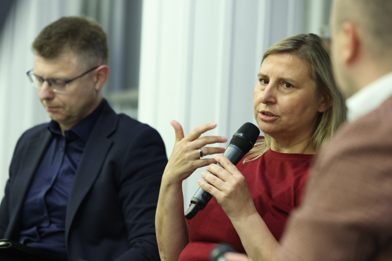 trzy osoby siedzą w sali jedna trzyma mikrofon 