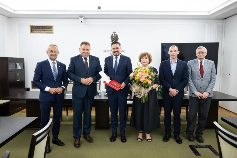 Sześć osób pozuje do zdjęcia stojąc w szeregu i uśmiechając się. W centrum stoi Marcin Wiącek trzymający w dłoniach okolicznościowy medal