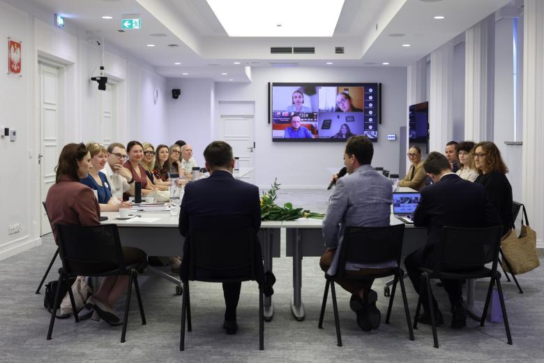 Kilkanaście osób siedzi przy stołach ustawionych w podkowę, na dużym telewizorze na ścianie widoczni uczestnicy posiedzenia łączący się zdalnie za pomocą programu do komunikowania się na odległość