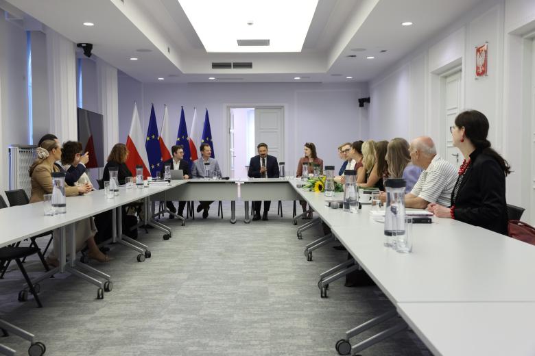 Kilkanaście osób siedzi przy stołach ustawionych w podkowę na sali konferencyjnej