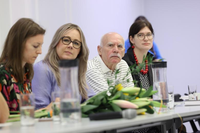 Cztery osoby siedzą przy stole w sali konferencyjnej i słuchają wystąpienia