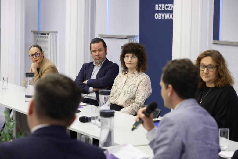 Sześć osób siedzi przy stole w sali konferencyjnej, jedna mowi do mikrofonu