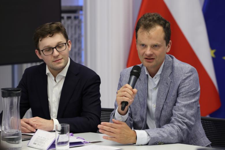 Dwie osoby siedzą za stołem w sali konferencyjnej, jedna z nich trzyma w dłoni mikrofon