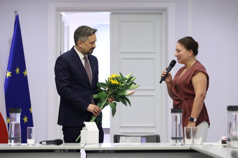 Dwie osoby stoją, jedna trzyma bukiet kwiatów, druga mówi do mikrofonu