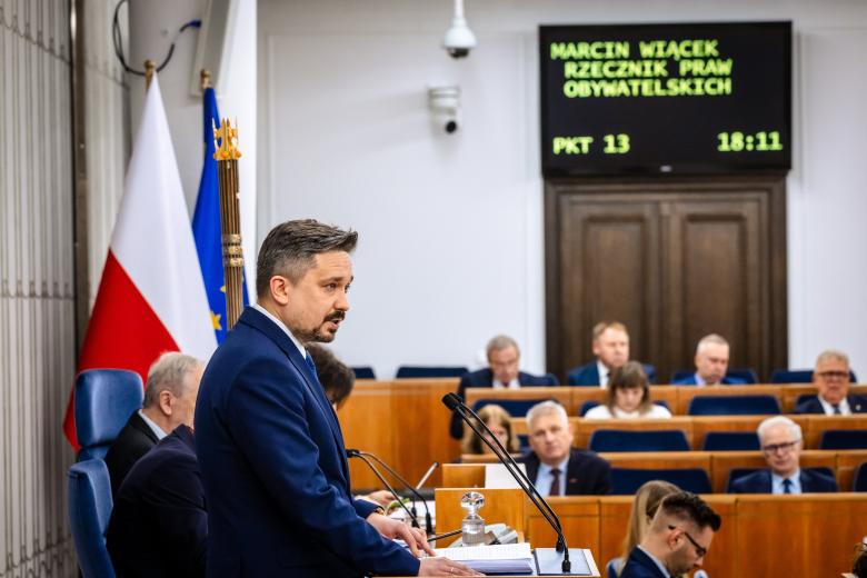 Osoba przemawia za mównicą na sali posiedzeń Senatu RP, wystąpieniu przysłuchują się osoby siedzące na sali obrad