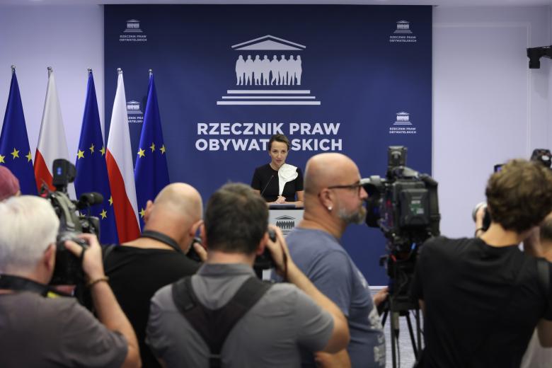 Osoba przemawia za mównicą, w tle duża granatowa plansza z napisem "Rzecznik Praw Obywatelskich" i flagi Polski i UE, na pierwszym planie stłoczeni fotoreporterzy i operatorzy z kamerami