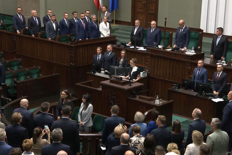 Osoba za mównicą sejmową odczytuje treść ślubowania, osoby obecne na sali obrad stoją