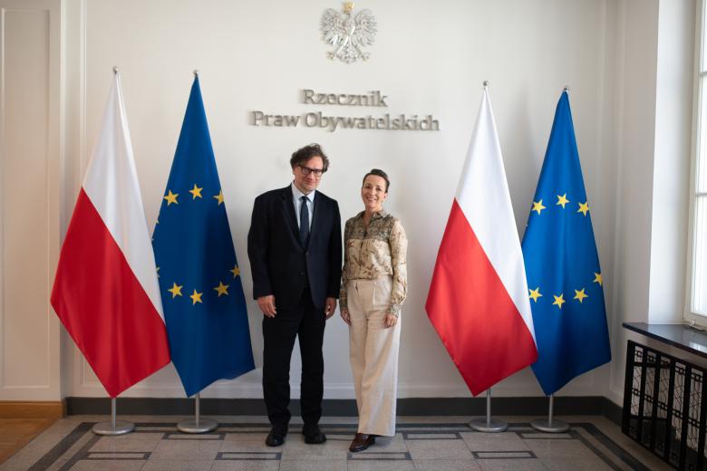 Dwie osoby pozują do wspólnego zdjęcia na tle flag Polski i UE i napisu "Rzecznik Praw Obywatelskich" na ścianie