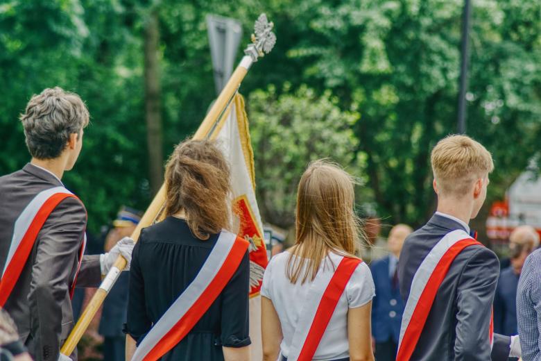 Czworo uczniów ubranych na galowo stoi trzymając sztandar szkoły podczas uroczystości szkolnej