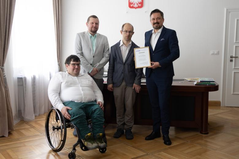 trzy osoby stoją w sali jedna siedzi na wózku 