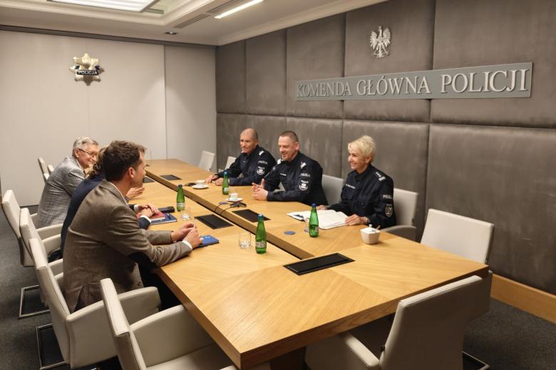 Sześć osób siedzi po dwóch stronach stołu konferencyjnego w sali z widocznym dużym napisem "Komenda Główna Policji" na ścianie