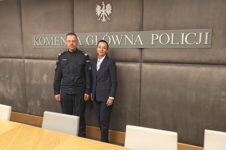 Dwie osoby pozują na stojąco do wspólnego zdjęcia na tle ściany z dużym napisem "Komenda Główna Policji"