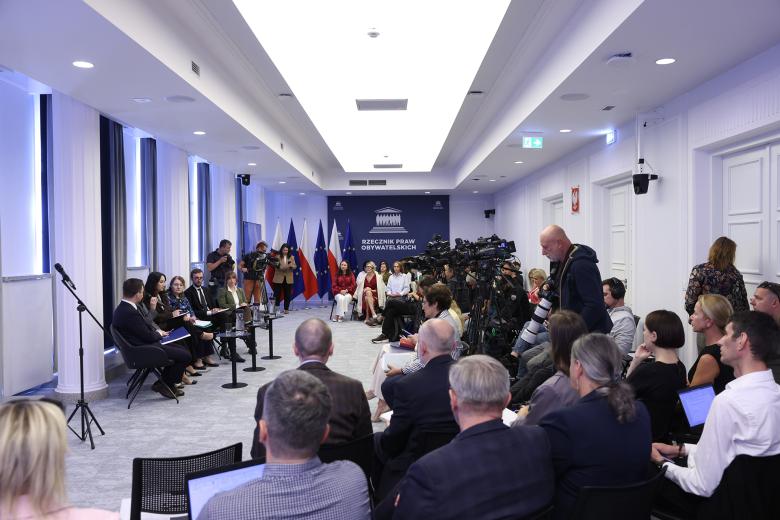 Widok ogólny sali konferencyjnej, na której siedzi kilkadziesiąt osób. Na środku sali sześć osób siedzi w rzędzie, naprzeciwko nich stoją kamery