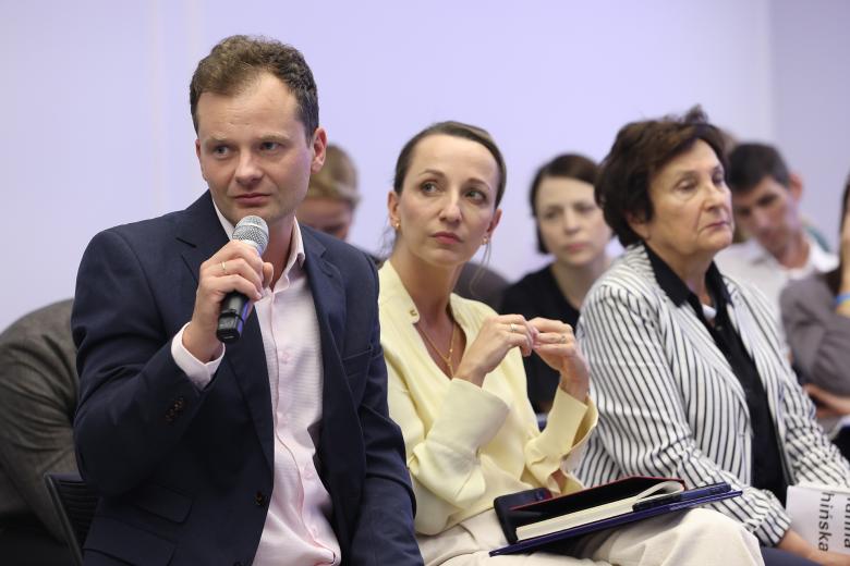 Adam Krzywoń siedzi i wypowiada się do trzymanego w dłoni mikrofonu. Obok niego siedzą RPO Sylwia Gregorczyk-Abram i dr Hanna Machińska
