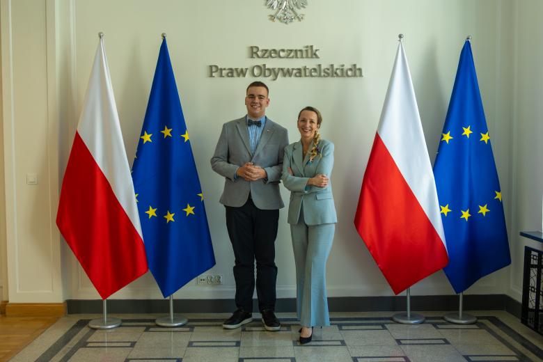 zdjęcie przedstawia dwie osoby pozujące do zdjęcia na tle flag PL i UE 