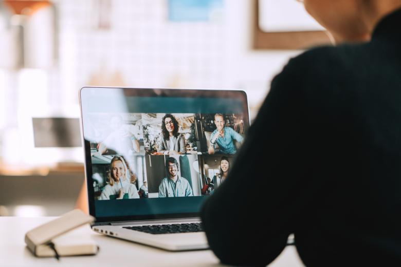 Osoba siedzi przy biurku, na którym stoi laptop z widocznym ekranem programu do przeprowadzania wieloosobowych spotkań w internecie