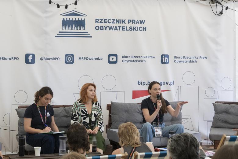 Trzy kobiety siedzą na kanapach przed publicznością. Jedna z nich wypowiada się do mikrofonu, gestykulując