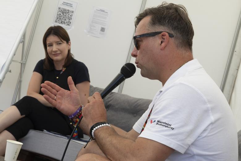 Mężczyzna siedzi na kanapie i wypowiada się do mikrofonu gestykulując. W tle siedzi przyglądająca się mu kobieta.