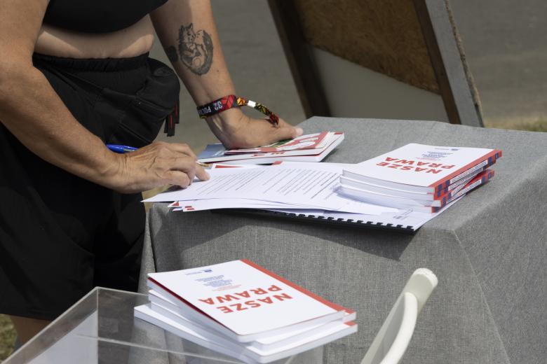 Kobieta stoi przy stoliku i wypełnia ankietę, w jednej z dłoni trzymając książkę i kilka ulotek. Na stole wyłożone są książki z tytułem "Nasze Prawa"