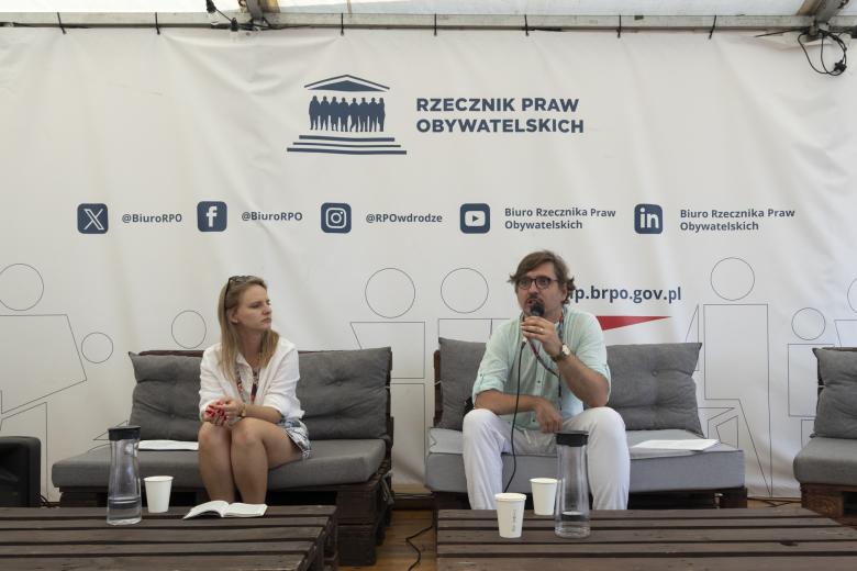 Mężczyzna i kobieta siedzą na kanapie w namiocie. Jedna z nich wypowiada się do mikrofonu