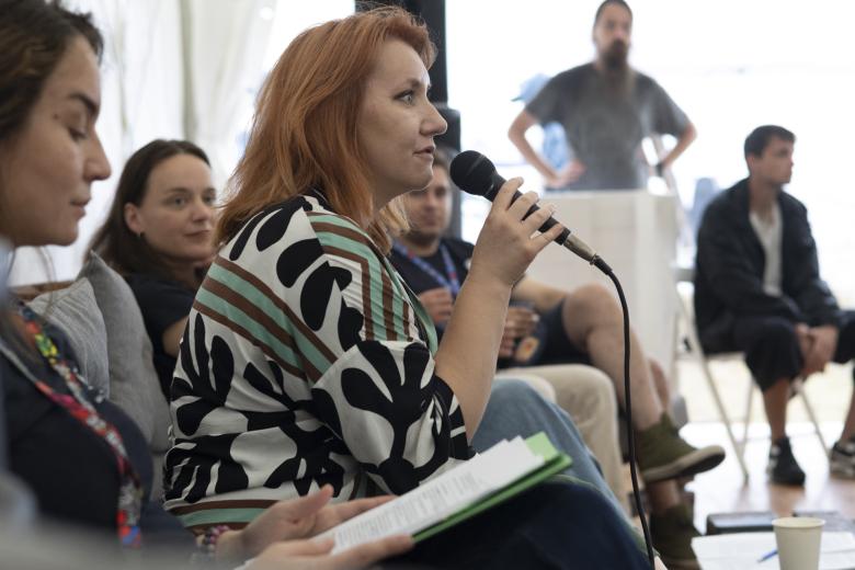 Trzy panelistki siedzą obok siebie na kanapie. Jedna z nich wypowiada się do mikrofonu. W tle widać osoby słuchające rozmowy