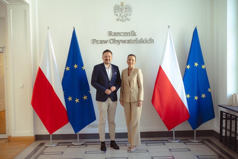 zdjęcie przedstawia dwie osoby - kobietę i mężczyznę - RPO obecnej iRPO poprzedniej kadencji pozujacych do zdjęcia na tle flag PL i UE oraz napisu Rzecznik Praw Obywatelskich
