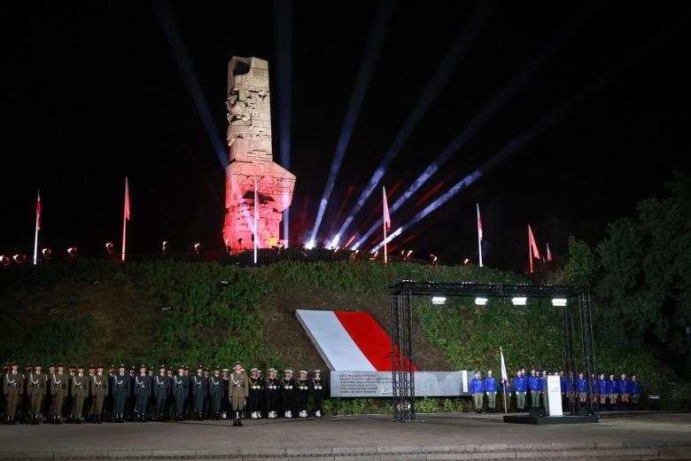 Uroczystości na Westerplatte - na kopcu stoi podświetlony biało-czerwono pomnik-obelisk otoczony masztami z flagami Polski. U stóp kopca w dwuszeregu stoi na baczność kilkadziesiąt żołnierzy.