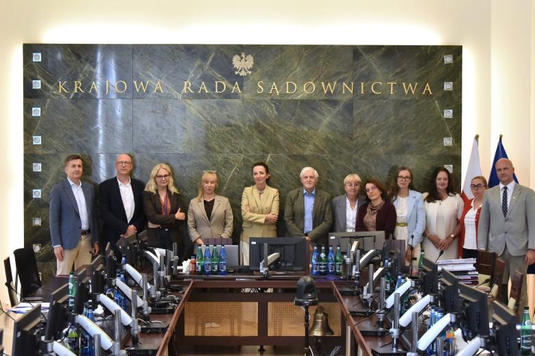 Dwanaście osób pozuje na stojąco do wspólnego zdjęcia na tle zielonej, marmurowej ściany ze złoconym napisem "Krajowa Rada Sądownictwa" i złoconym orłem w koronie