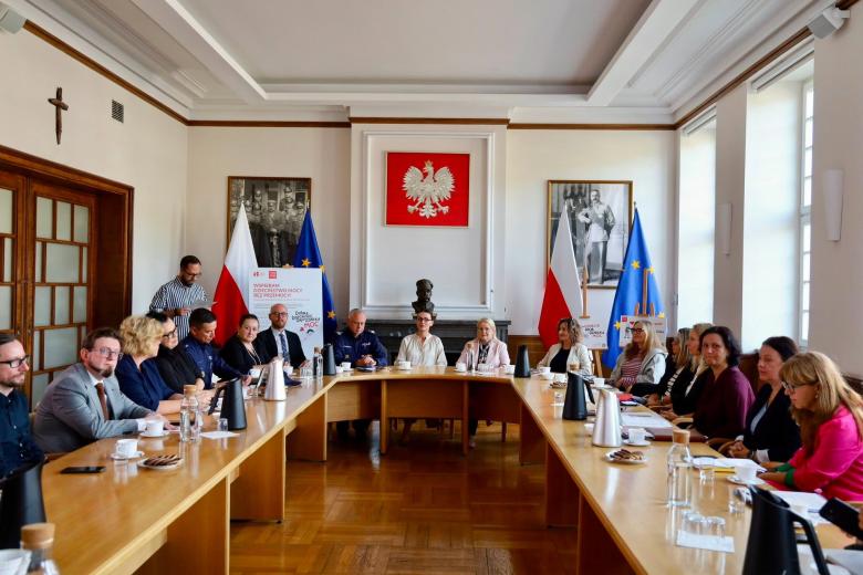 Kilkanaście osób siedzi przy dużym stole ustawionym w podkowę w sali narad urzędu z dużym godłem Polski na ścianie i flagami Polski i UE.