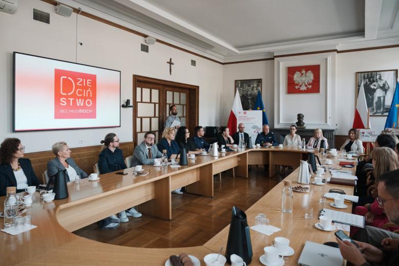 Kilkanaście osób siedzi przy dużym stole w sali narad urzędu z dużym godłem Polski na ścianie i flagami Polski i UE, a z boku z wyświetlaczem, na którym znajduje się logo kampanii z napisem "Dzieciństwo bez Przemocy"