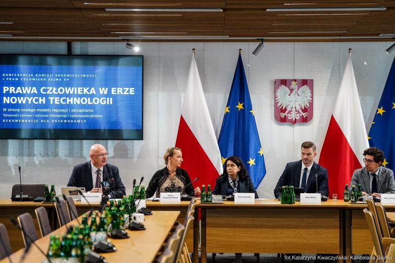 Pięć osób siedzi za stołem na sali konferencyjnej, za nimi flagi Polski i UE oraz wiszące na ścianie godło Polski