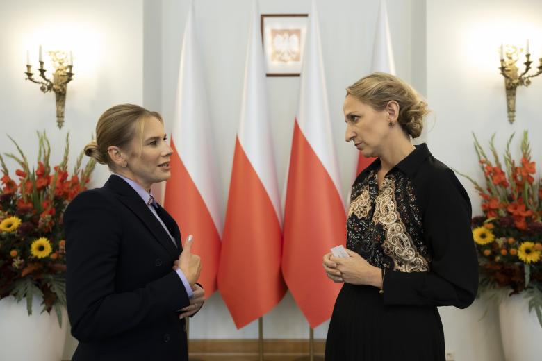 Dwie osoby rozmawiają w pomieszczeniu na tle flag Polski
