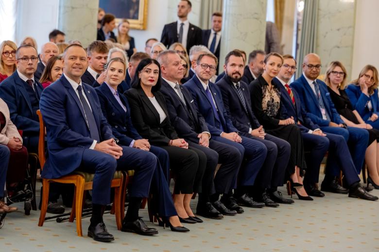 Zbliżenie na grupę osób siedzącą w pierwszym rzędzie krzeseł na dużej sali i słuchających czyjegoś wystąpienia