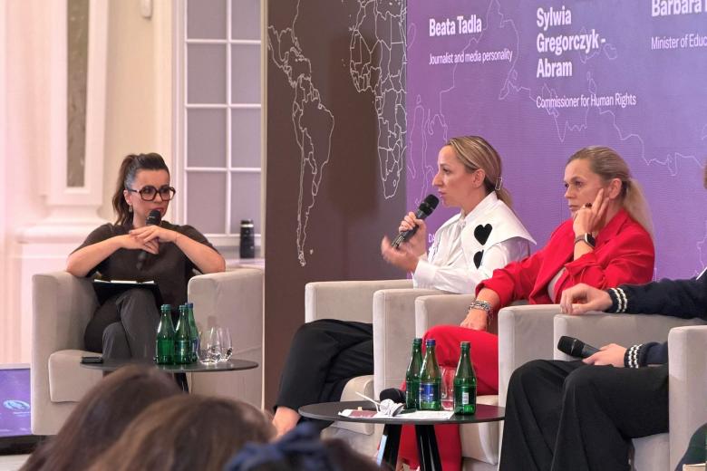 Trzy osoby siedzą w fotelach na scenie, jedna mówi do trzymanego w ręku mikrofonu