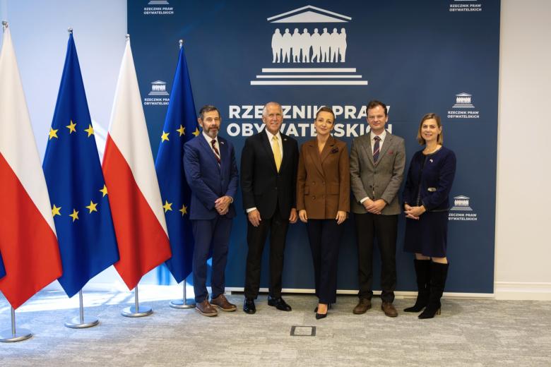 Pięć osób pozuje na stojąco do wspólnego zdjęcia na tle ścianki z napisem "Rzecznik Praw Obywatelskich" i flag Polski i UE