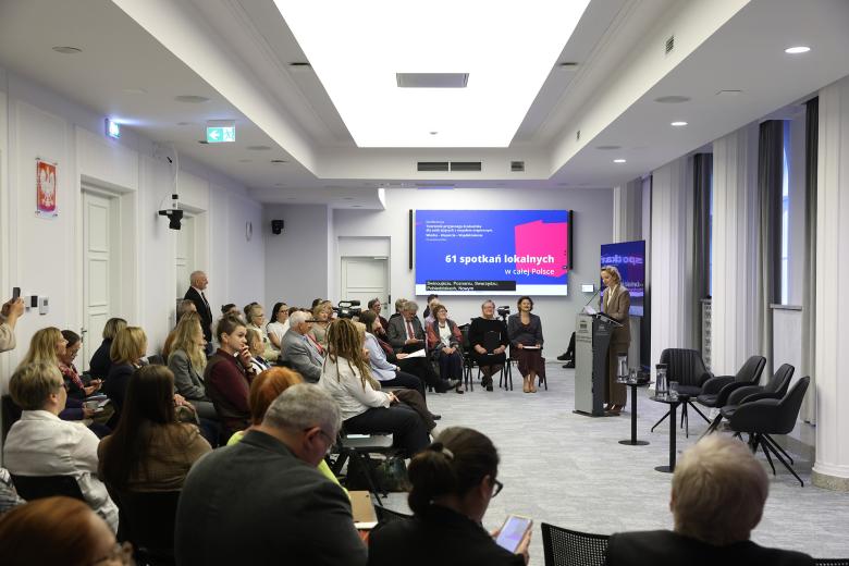 RPO Sylwia Gregorczyk Abram stoi przy mównicy i wypowiada się. Naprzeciwko jej na widowni siedzi kilkadziesiąt osób. W tle na wyświetlaczu grafika z napisem "61 spotkań lokalnych"